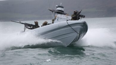 The Commando Raiding Craft is seen in action in Plymouth Sound. It's seen moving in a coastal body of water, with at least six personnel on the craft. Two soldiers are manning what appears to be machine guns, one in front of the boat and the other in the back. In the background, a coastal land mass is seen amid foggy weather.