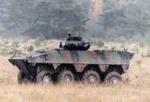 The French Army's VBCI Infantry Fighting Vehicle is seen moving across a brown, grassy area. The vehicle is painted in green, black, and brown camouflage. The background is a forested area.