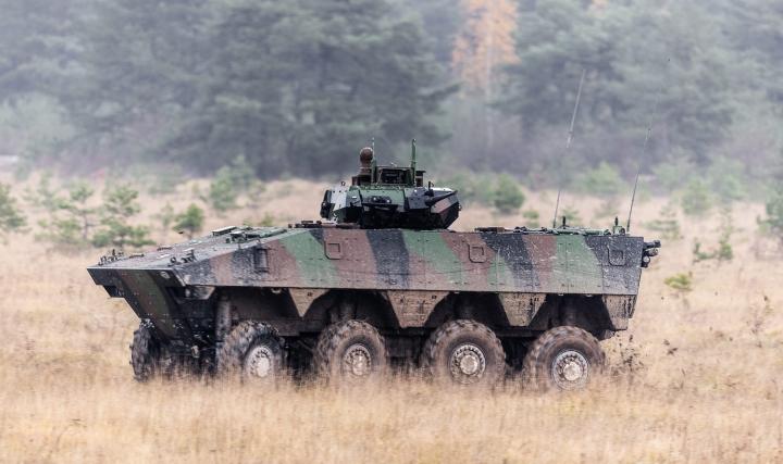 The French Army's VBCI Infantry Fighting Vehicle is seen moving across a brown, grassy area. The vehicle is painted in green, black, and brown camouflage. The background is a forested area.