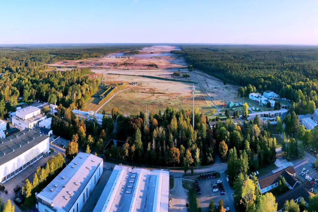 A view of the landscape where Werk Niedersachsen will be constructed. The foreground of the image is a complex made up of several buildings. Behind them, a forested area is shown, its middle stripped off of trees and its brown land flattened. The area stretches over the horizon.