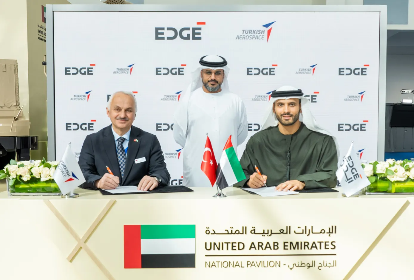 Turkish Aerospace President and CEO Professor Temel Kotil and EDGE Group Platforms & Systems President Khaled Al Zaabi are seen signing documents during a memorandum of agreement ceremony. In the back, a facilitator oversees the procedure. A white panel with EDGE and Turkish Aerospace's logos printed on it serves as their background. The table where the signing is taking place on has the UAE, Turkey, and the company's flags propped up on it.