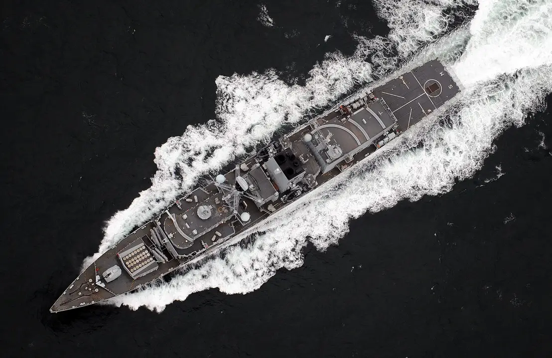 A UK Royal Navy ship is seen sailing deep dark-blue waters. The scene is shown from a top-down view.