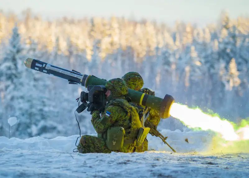 Two camouflaged soldiers fire off a Javelin anti-tank missile pointing northwest. The launcher is seen emitting yellow-green flames from its back. The scene is taking place in a snowy area, with a forest of snow-covered trees blurred out in the back.