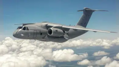 An Embraer C-390 Millennium aircraft is seen flying above the clouds. The gray aircraft has the words "HUNGARIAN AIR FORCE" painted on the side of the cockpit in black. The number 610 is also painted on the jet engine, the wing, and the side of the cockpit, just below one of the side windows. The background is a bright blue sky.