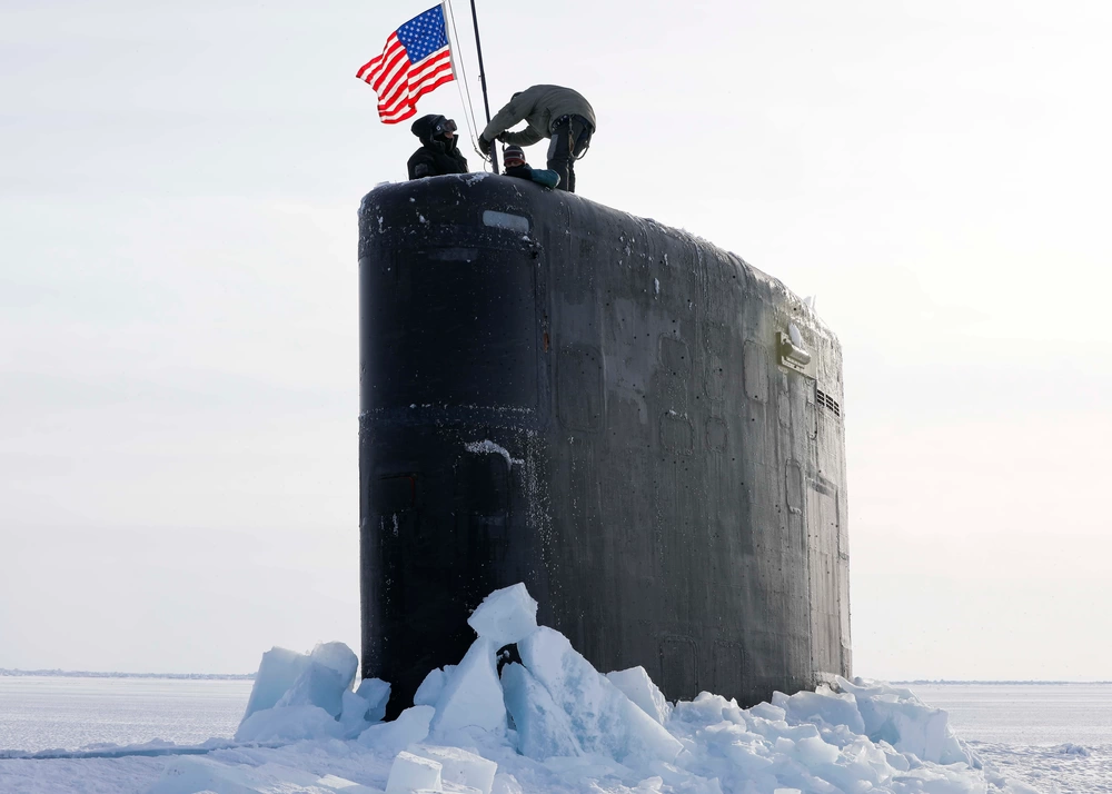 The USS Hampton's sail is seen breaking through the Arctic's massive ice flats. The submarine's sail is the only visible part of the vehicle. The ship is painted black. On top of the sail, three individuals are attaching a flag of the US on what appears to be a makeshift flag pole. The background is a completely white landscape and sky blending seamlessly together.