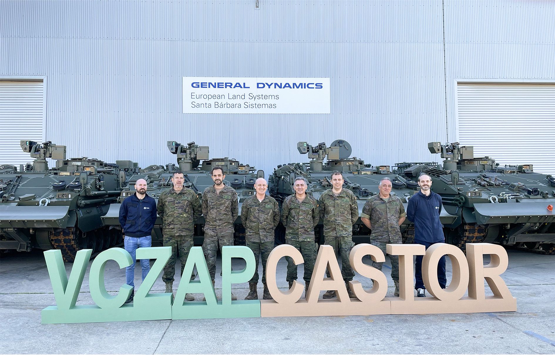 Six uniformed Spanish Army personnel stand in front of four ASCOD Castor armored combat vehicles. They are joined by two civilians, likely from General Dynamics. In front of them are letter standees that spell out "VCZAP CASTOR". The background is a warehouse building, with a sign that reads "GENERAL DYNAMICS European Land Systems Santa Barbara Sistemas".