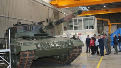 A Leopard 2A4 main battle tank is seen inside a warehouse facility, where 15 onlookers stand by its left side while inspecting it. The tank is painted in camouflage colors.