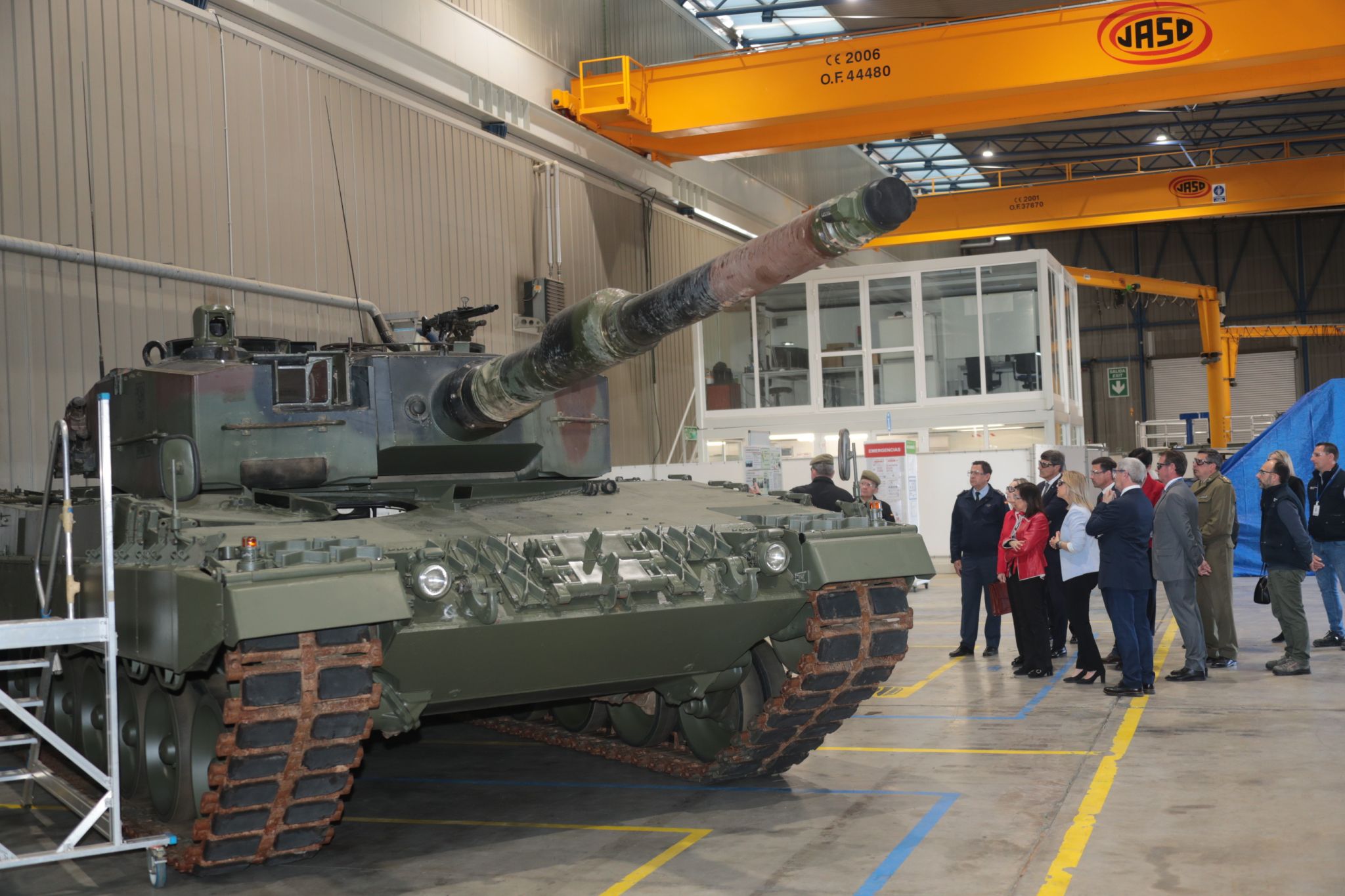 A Leopard 2A4 main battle tank is seen inside a warehouse facility, where 15 onlookers stand by its left side while inspecting it. The tank is painted in camouflage colors.