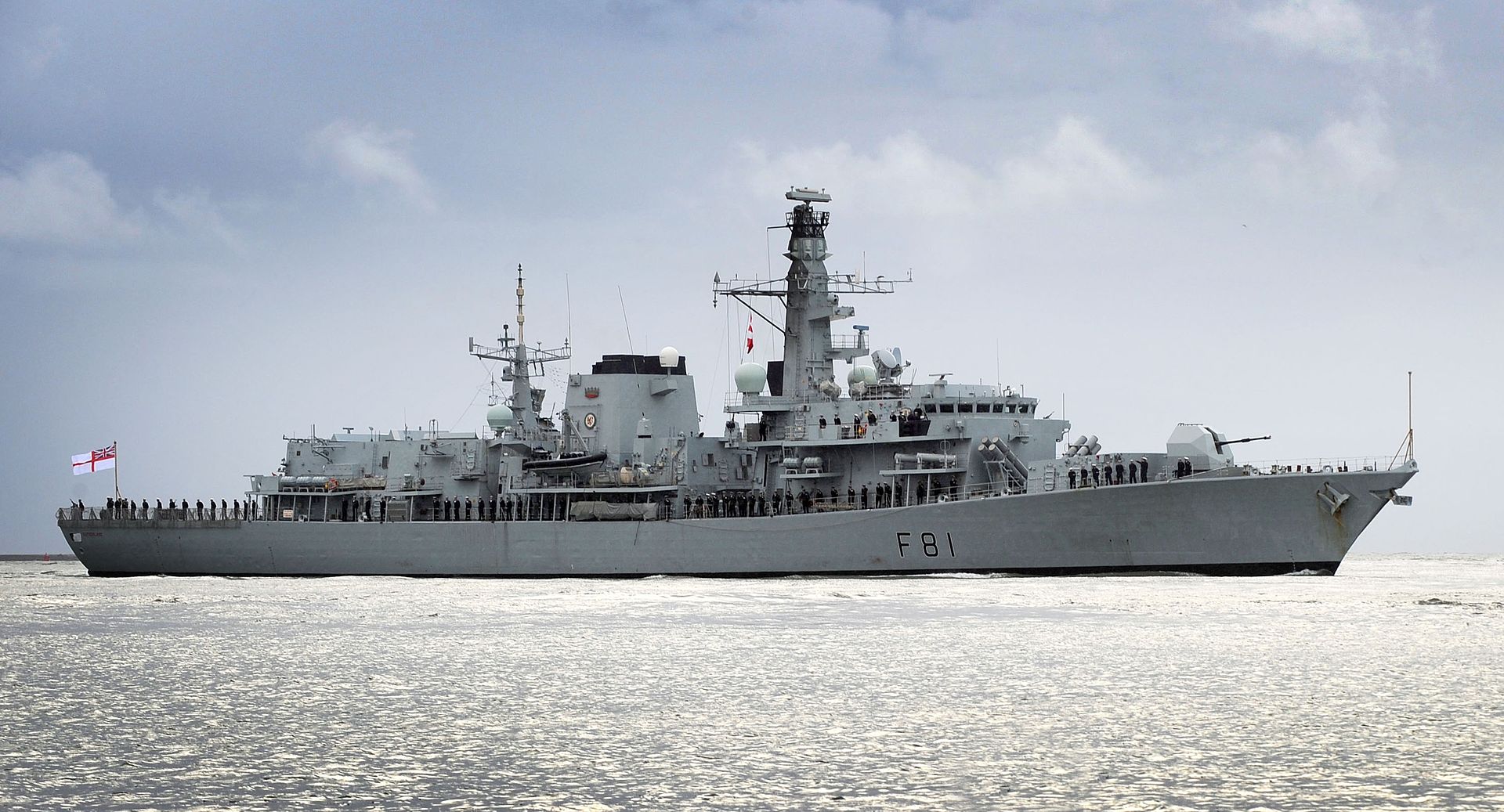 HMS Sutherland, a UK Royal Navy frigate, is seen sailing on calm waters. The gray ship has "F81" painted in black on its side. Around 60 of its crew members are seen standing on deck, wearing their navy blue uniforms and white sailor hats. The sky is shrouded in light gray clouds.
