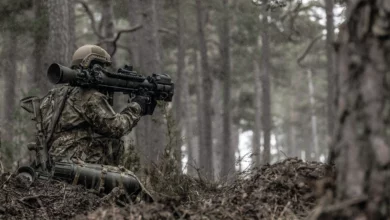 A soldier is seen with his back turned, seemingly laying low in a brown, forested area. He is wearing camouflage. On his right shoulder, a black Carl-Gustaf rifle rests as he props it up to point forward.