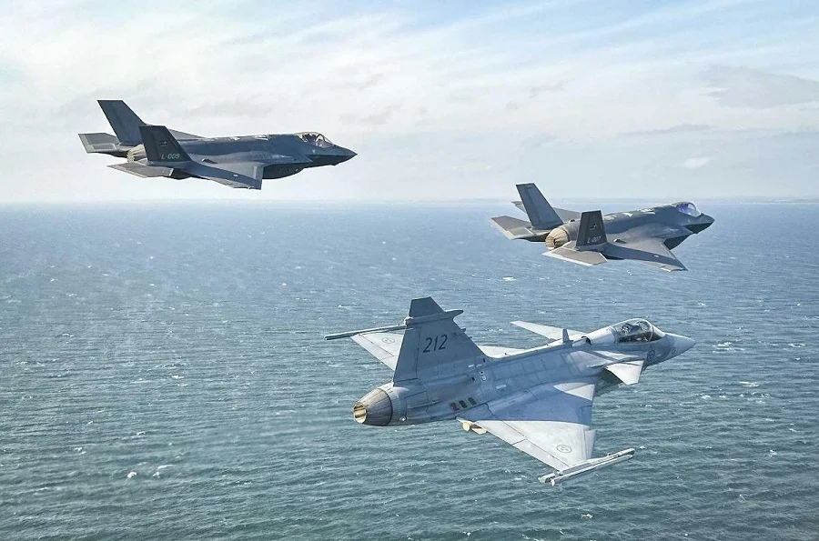 Two F-35 Lightning II and a JAS-39 Gripen fighter plane fly together during Denmark and Sweden's drills. The background is a clear blue sky with some white clouds covering it. The sea below shows calm, blue-green waters.