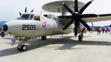 A Northrop Grumman E-2K Hawkeye plane appears to be on display at a wide-open area similar to a runway. It is cordoned off by a red-velvet stanchions seen in the back. The gray plane has "2505" painted on its left side in black, below the cockpit. It also has a red-white-blue logo with an eagle's head. Above it's body, its circular radar system is seen set parallel to its body.