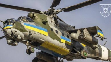 A Mi-24 helicopter is seen flying overhead. The helicopter is painted in camouflage, with stripes of the Ukrainian flag's blue and yellow colors running its body horizontally.
