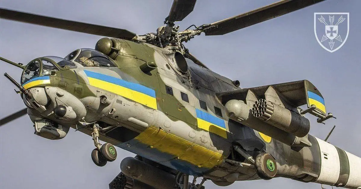 A Mi-24 helicopter is seen flying overhead. The helicopter is painted in camouflage, with stripes of the Ukrainian flag's blue and yellow colors running its body horizontally.