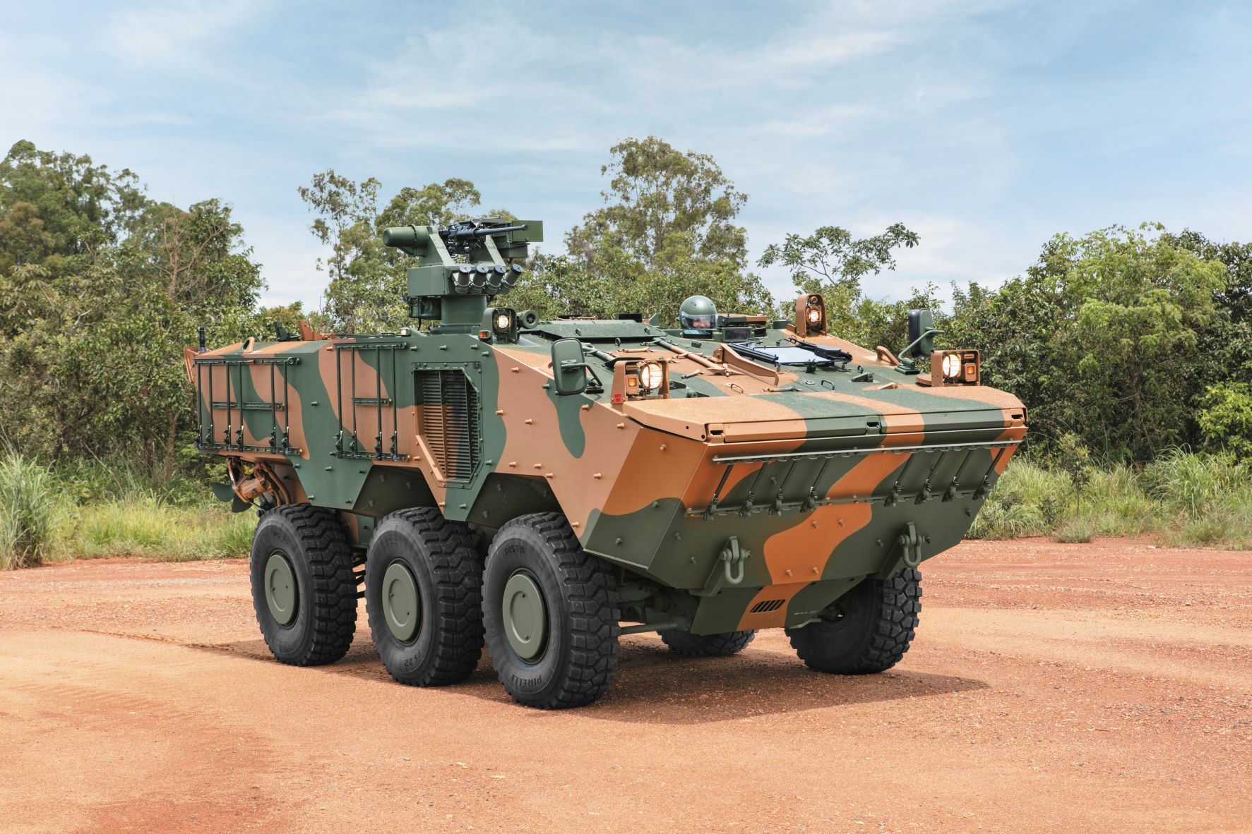 A Guarani 6x6 Armored Personnel Carrier is seen parked in an area with red sand and some trees and bushes in the background. The vehicle is painted in green and reddish-brown camouflage. A soldier wearing a helmet is seen peeking out the hatch. The far background is a blue sky with thin white clouds scattered evenly.