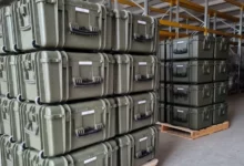 Dozens of military equipment boxes are seen stacked on top of each other in what appears to be a warehouse. They are supported underneath by wooden crates.