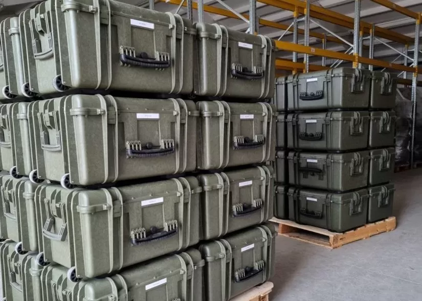 Dozens of military equipment boxes are seen stacked on top of each other in what appears to be a warehouse. They are supported underneath by wooden crates.
