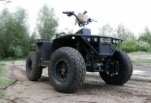 A REEQ Rex Quad is seen parked in a muddy field with trees and shrubbery in the background. The vehicle is a 4-wheel off-road vehicle with large wheels and a minimalistic chassis. Most of its parts are painted black, except for the logo in front and the casing of the screen in the middle of the handlebars, which are both dirty brown.