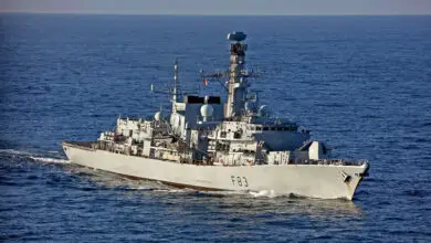 The UK Royal Navy's HMS St Albans frigate is seen sailing in calm, deep blue waters. The ship is painted gray, with "F83" painted on the right side of its hull in black. The far background is a gray sky.