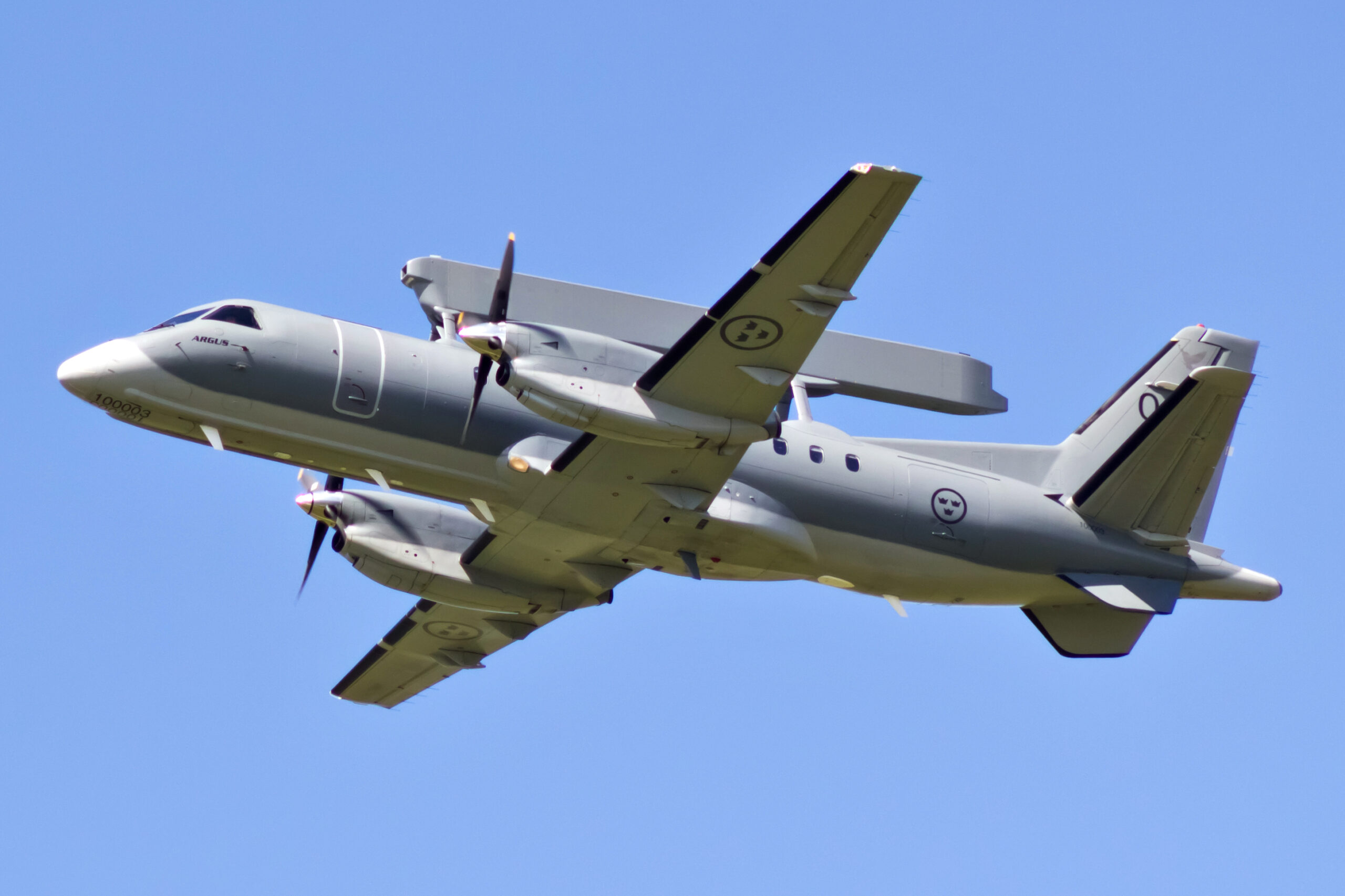 A Saab 340 aerial early warning and control plane is seen flying in clear blue skies. Its long, rectangular radar system is seen connected and propped up parallel to its body. The aircraft is painted gray. The photo was taken underneath the plane.