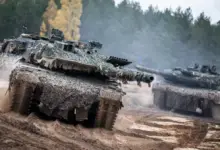 Three armored tanks are seen moving in a sandy area with trees in the back. The tanks are covered with ghillie suit fabric to help with camouflaging.