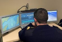 A uniformed French Navy personnel has his back turned against the camera as he sits in front of a computer system with three computer screens propped up on a table. The screens show what appears to be the interface of Thales' Expeditionary Portable Operations Centre (e-POC). The left screen displays a topographic map, and the middle screen shows various data through lines and graphs. The screen on the right appears to be displaying a blue-violet wallpaper.