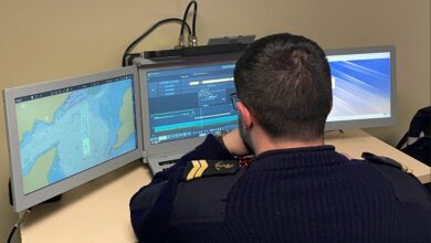 A uniformed French Navy personnel has his back turned against the camera as he sits in front of a computer system with three computer screens propped up on a table. The screens show what appears to be the interface of Thales' Expeditionary Portable Operations Centre (e-POC). The left screen displays a topographic map, and the middle screen shows various data through lines and graphs. The screen on the right appears to be displaying a blue-violet wallpaper.