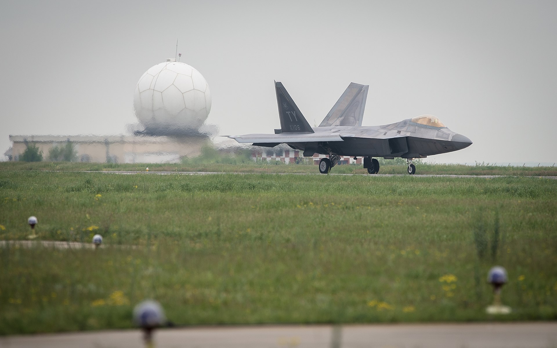 A gray F-22A Raptor jet plane taxis at Mihail Kogalniceanu Airport, with a globe-shaped radar station radome visible in the background.