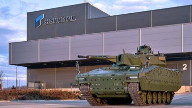 A green armored tracked tank is seen parked outside of a Rheinmetall facility. The building in the back has the Rheinmetall logo in front, attached on the upper left side of the facade. Some red-brown shrubbery is seen on the middle left side of the image, planted in some partition area between the parking lot and the building. The sky in the background is a blue one with some white and gray clouds interspersed.