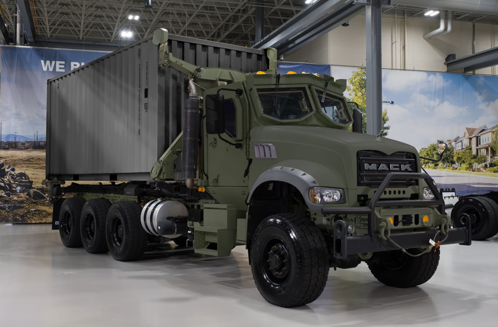 A Mack Common Tactical Truck is seen on display at what appears to be an arms/miitary exhibit. The 8-wheel truck is painted deep green. It has a cargo container in the back, painted a dull gray.