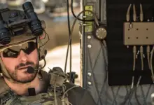 A soldier is seen beside INVISIO's Intercom system, a rectangular device with eight ports on its top and bottom sides. The soldier, equipped in camouflage, also has a headset, a pair of binoculars, and a radio.