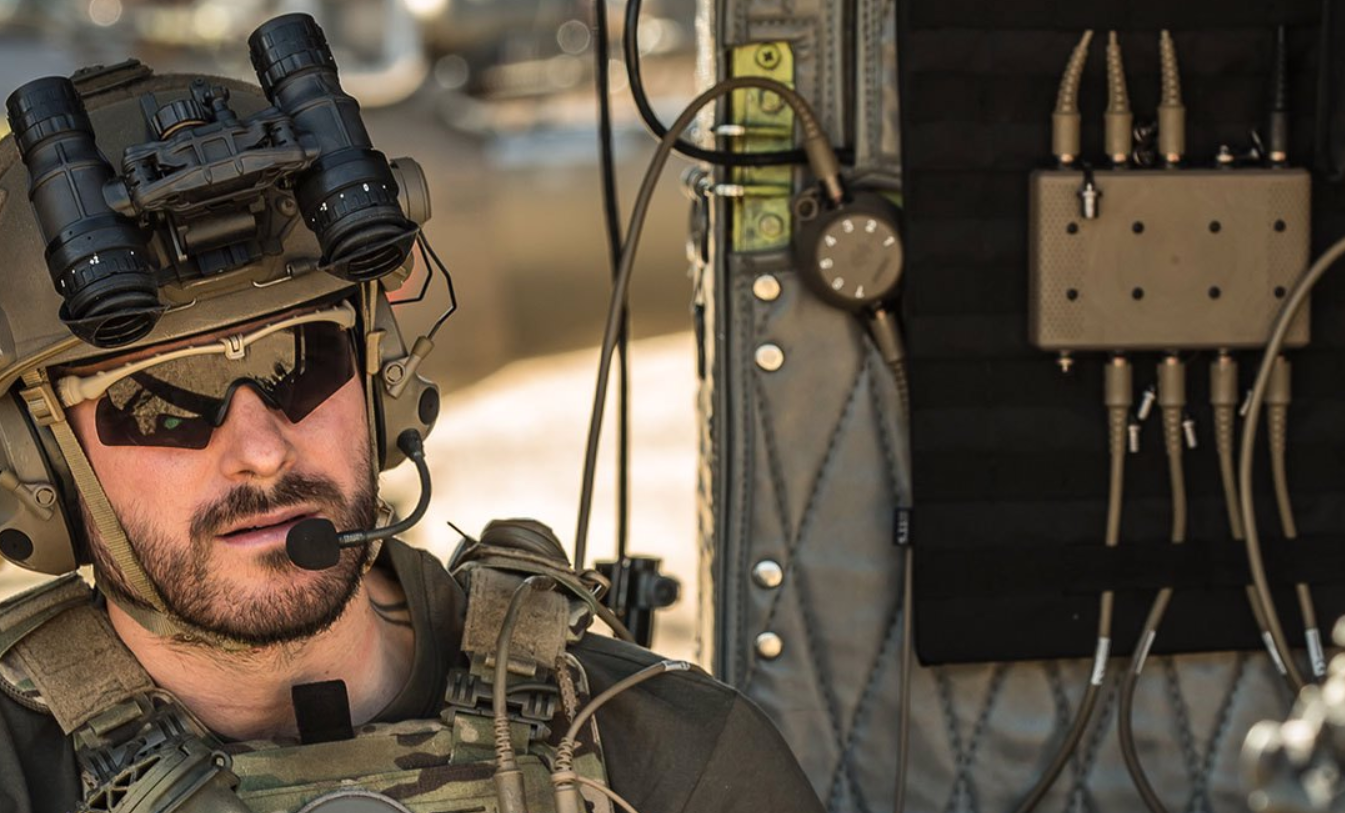 A soldier is seen beside INVISIO's Intercom system, a rectangular device with eight ports on its top and bottom sides. The soldier, equipped in camouflage, also has a headset, a pair of binoculars, and a radio.