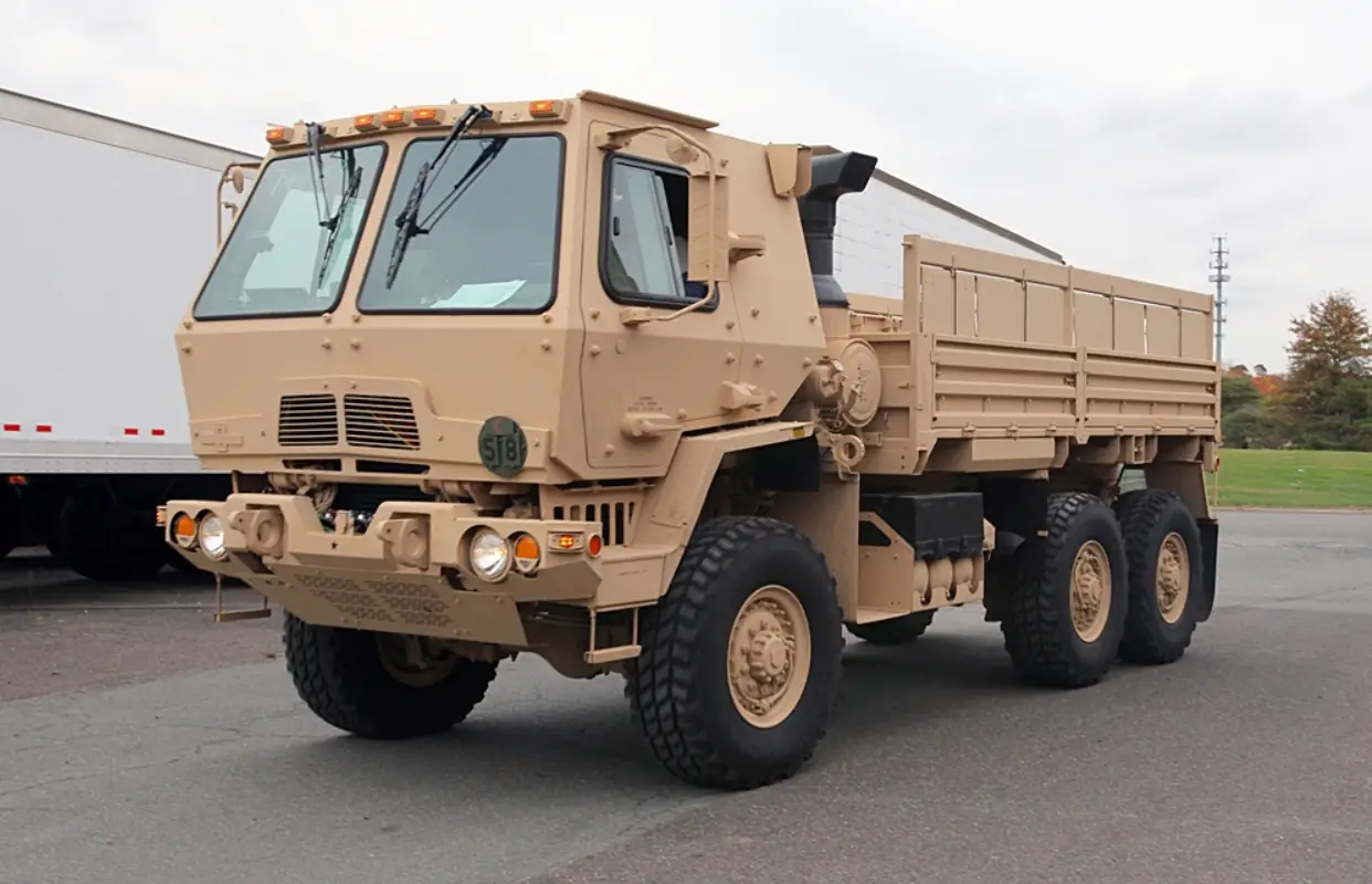 A brown M1083 army truck is seen parked in what appears to be a parking area. The six-wheel truck has two windshields and an open-trunk back.