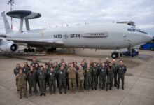 The women of the NATO Airborne Early Warning and Control Force pose for a photo in front of a NATO AWACS plane. They appear to be on a plane runway. The soldiers are all wearing green fatigue overalls. The sky in the background is covered by white heavy clouds.