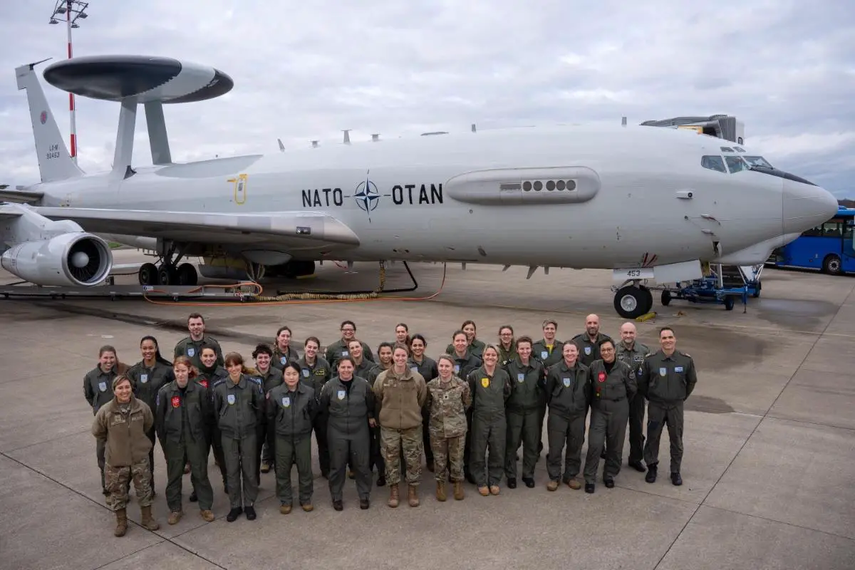 The women of the NATO Airborne Early Warning and Control Force pose for a photo in front of a NATO AWACS plane. They appear to be on a plane runway. The soldiers are all wearing green fatigue overalls. The sky in the background is covered by white heavy clouds.