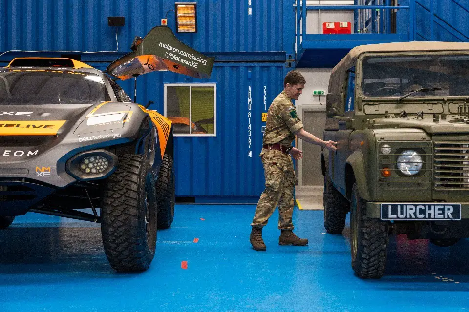 A British Army land rover with an electric engine is seen parked on the right side of the image, right beside a NEOM McLaren Extreme E racecar on the left. A uniformed army personnel is seen reaching for the land rover's door, as if opening it. The rover is painted brown-green, with a plate in its front that reads "LURCHER".The racecar is painted black with orange decals.