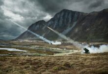 Two National Advanced Surface to Air Missile Systems (NASAMS) are seen firing off missiles from a grassy field in front of a mountainous landscape. In the far back, the sky is covered by dark and heavy clouds.