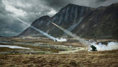Two National Advanced Surface to Air Missile Systems (NASAMS) are seen firing off missiles from a grassy field in front of a mountainous landscape. In the far back, the sky is covered by dark and heavy clouds.