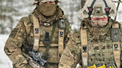 Two camouflaged soldiers appear to be wearing Saab's live training equipment, which tracks the soldiers during training with laser and geometric pairing technology. Various black straps and sensors can be seen attached to their helmets.