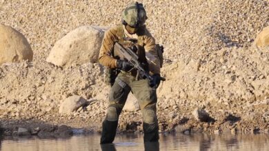 A soldier standing on shallow waters is seen wearing a helmet and fatigues and holding a Steyr GL-40 grenade launcher. The weapon is a portable capability smaller than an automatic rifle. The background is the water's shores made up of light brown sand and rock.