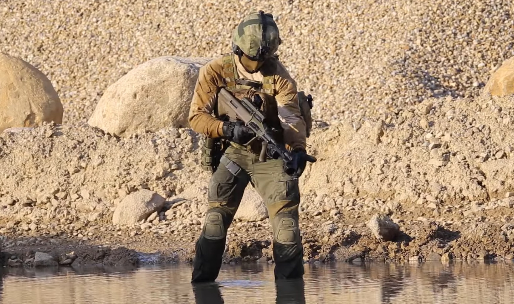 A soldier standing on shallow waters is seen wearing a helmet and fatigues and holding a Steyr GL-40 grenade launcher. The weapon is a portable capability smaller than an automatic rifle. The background is the water's shores made up of light brown sand and rock.