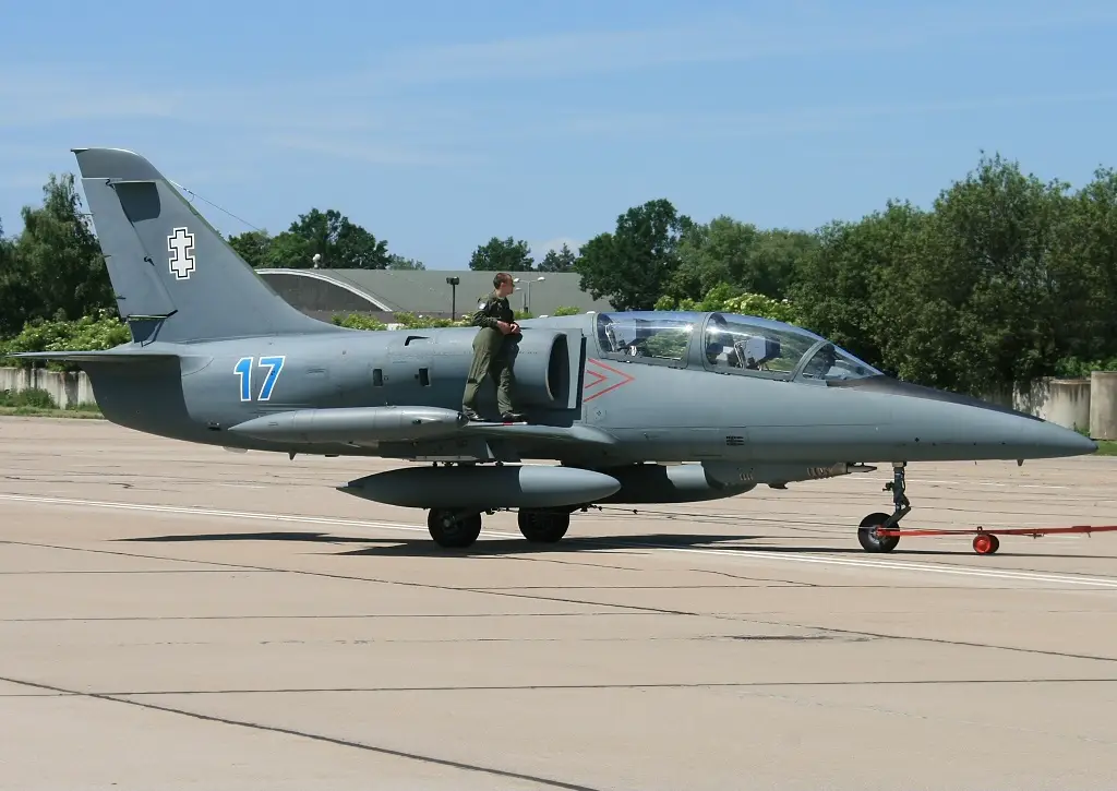 A Lithuanian Air Force Aero L-39ZA Albatros is seen parked in a wide-open cemented field. The gray aircraft has the number 17 painted on its side, by the tail. A soldier wearing green fatigues is seen standing on its wing and leaning on its body. The background is a tree-filled area with a clear blue sky farther in the back.