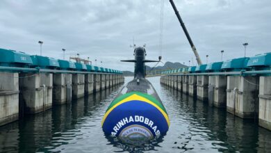 The Tonelero (S42), Brazil's third Riachuelo-class submarine, is seen half-submerged in a pier. Its front is wrapped in clothing with Brazil's colors, as well as the Brazilian Navy's logo in the center. The background is a dull sky covered in dark clouds.