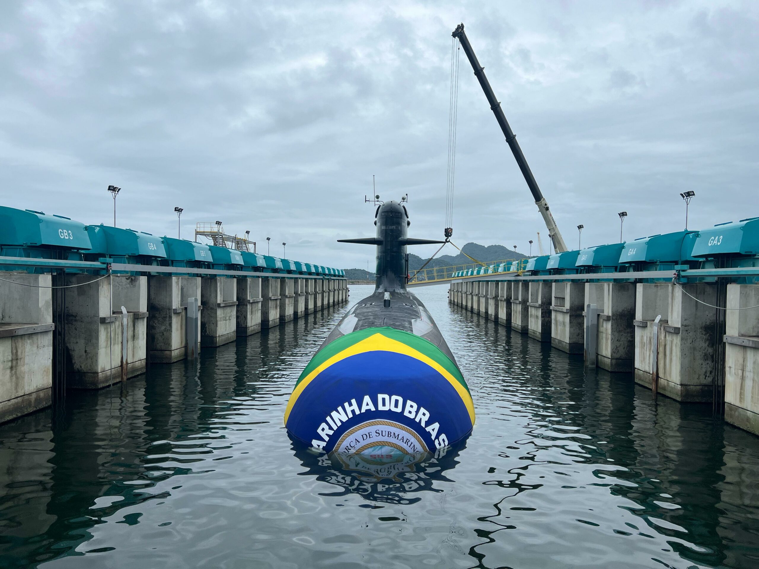 The Tonelero (S42), Brazil's third Riachuelo-class submarine, is seen half-submerged in a pier. Its front is wrapped in clothing with Brazil's colors, as well as the Brazilian Navy's logo in the center. The background is a dull sky covered in dark clouds.