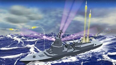 An artist's impression of the AN/SPY-7(V)1 radar-fitted Aegis System Equipped Vessel (ASEV). The gray ship is seen sailing in rough waters. Three missiles are seen firing off from it, with one maneuvering to hit an airborne missile fired by an unknown source. By the back of the ship, three laser-like lights target a group of five drones flying above. Two triangle-shaped objects are seen hovering above the entire scene.