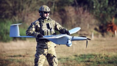 A uniformed soldier holding a WB Group FlyEye unmanned aerial vehicle in a grassy area with trees and shrubbery in the back. The white lightweight drone has a shape similar to high wing aircraft, with its wings attached on top of its body.