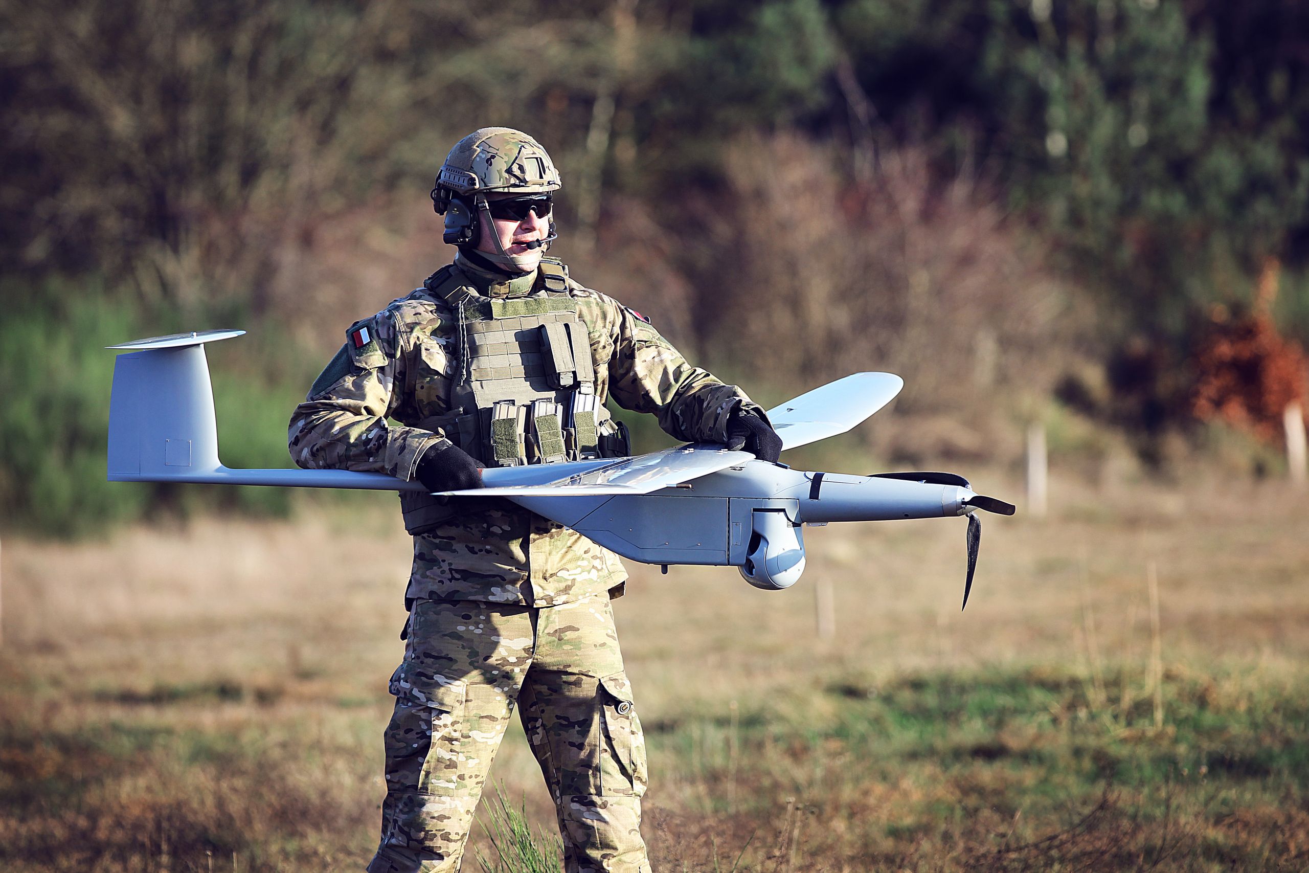 A uniformed soldier holding a WB Group FlyEye unmanned aerial vehicle in a grassy area with trees and shrubbery in the back. The white lightweight drone has a shape similar to high wing aircraft, with its wings attached on top of its body.