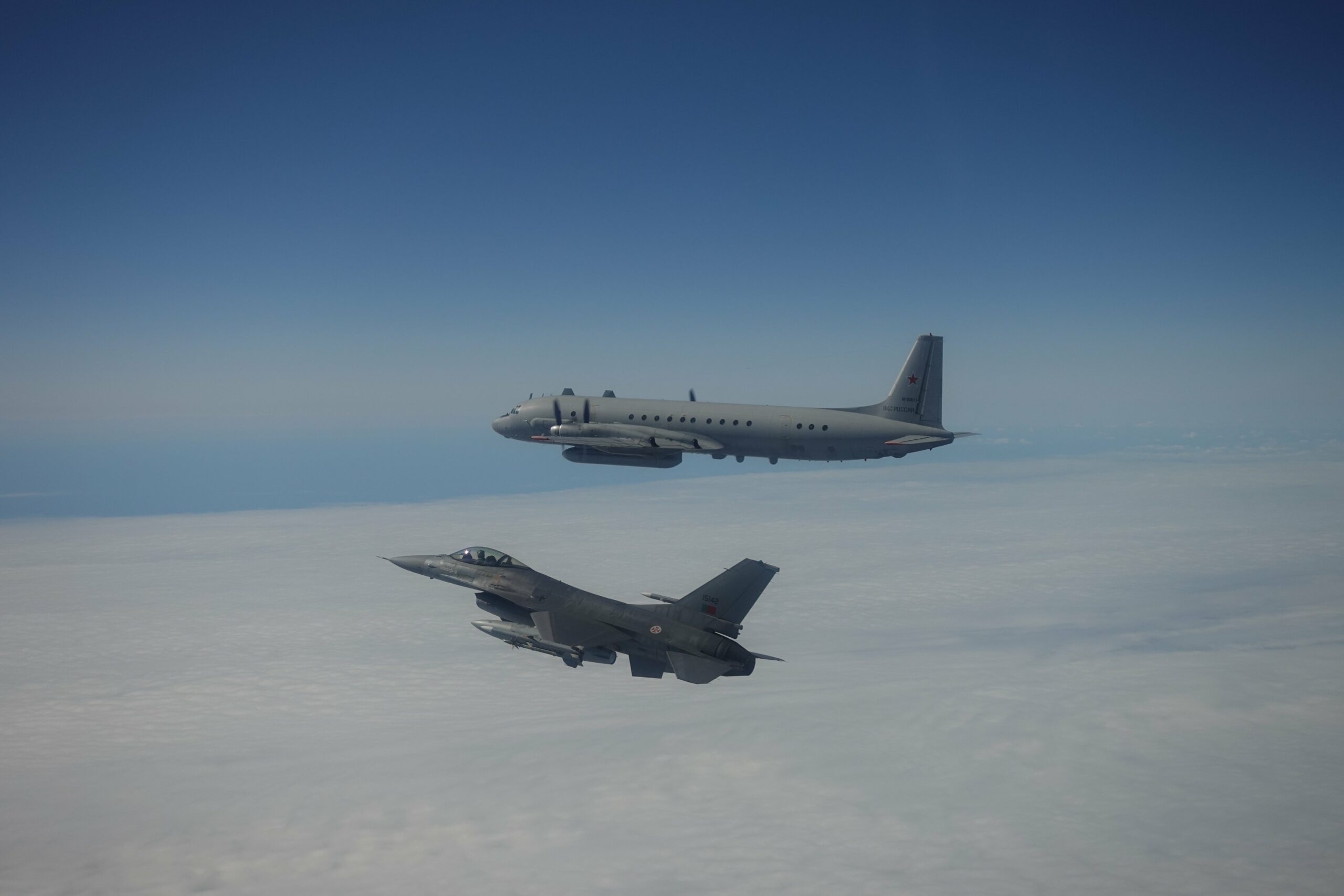 A Portuguese F-16 fighter jet escorting a Russian aircraft out of NATO's airspace above the clouds. Both planes are painted gray. The background is a sea of white clouds and the clear blue sky above.