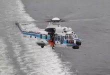 A Japan Coast Guard H225 helicopter is seen flying above gray waters. The white aircraft has a thin navy blue and thick light blue stripe painted on its side, as well as "JAPAN COAST GUARD" written on its tail boom. Three personnel can be seen inside the helicopter: one is the pilot inside the cockpit, and the other two are on the passenger seats.