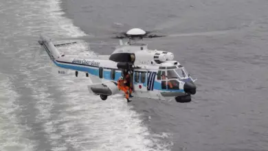 A Japan Coast Guard H225 helicopter is seen flying above gray waters. The white aircraft has a thin navy blue and thick light blue stripe painted on its side, as well as "JAPAN COAST GUARD" written on its tail boom. Three personnel can be seen inside the helicopter: one is the pilot inside the cockpit, and the other two are on the passenger seats.
